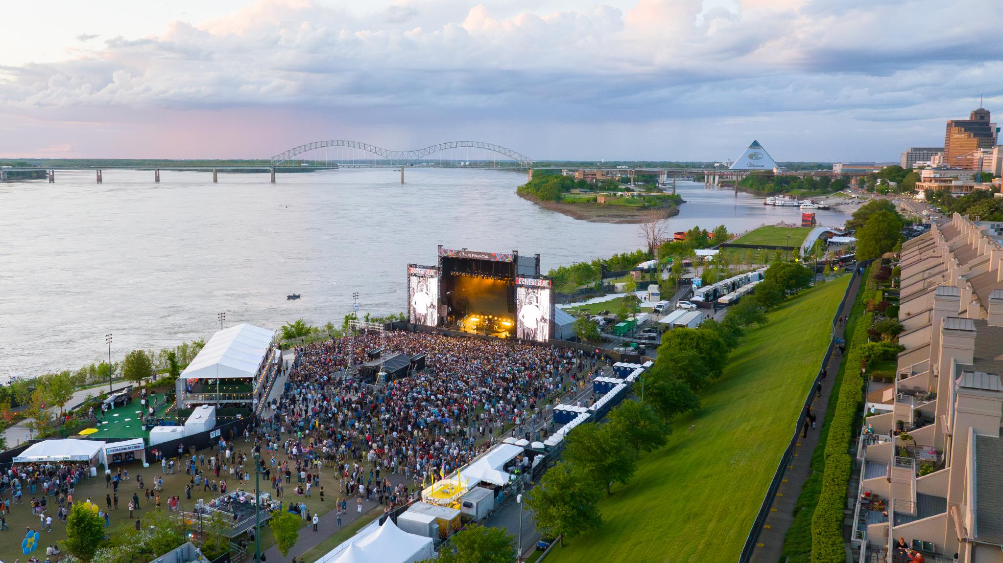 A crowd gathering at a stage for RiverBeat Music Festival at Tom Lee Park, with the Mississippi River, the downtown Memphis skyline and Hernando de Soto Bridge in the background.