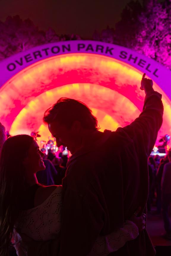 Couple listens to a concert at Overton Park Shell, which is illuminated in pink and orange neon lights.