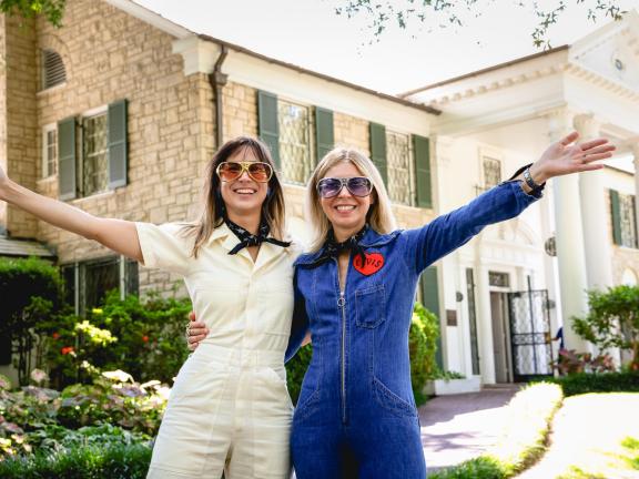two women in jumpsuits pose in front of Graceland mansion