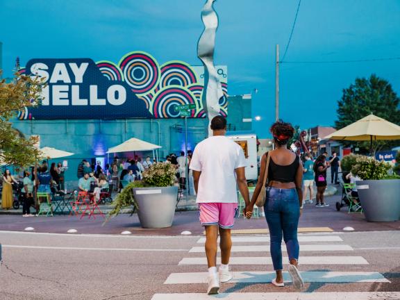 couple holding hands walks across the street in front of blue wall with say hello sign