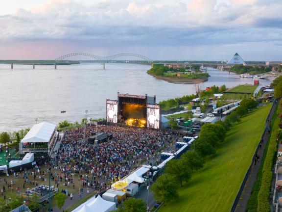 An aerial view of RiverBeat Music Fest, with a large crowd gathered in front of a large stage. The Mississippi River, Hernando de Soto Bridge and Memphis skyline are in the background.