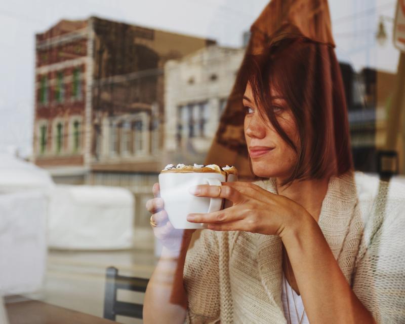 Photo from outside of window of a woman drinking coffee with reflection of buildings showing