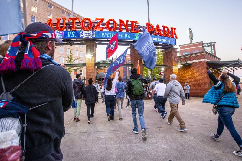 901 FC soccer fans walk into AutoZone Park below the stadium sign carrying blue and red flags