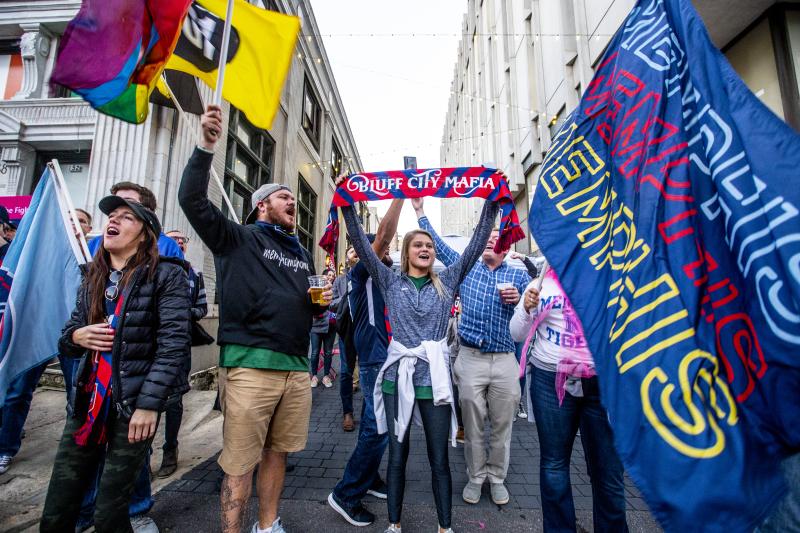 901 fc fans stand cheering in an alley during the march prior to the game. they're holding flags that say Memphis and Bluff City Mafia