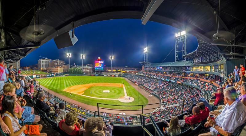 Panoramic photo of baseball stadium full of fans in the evening. The diamond is lit up by the lights