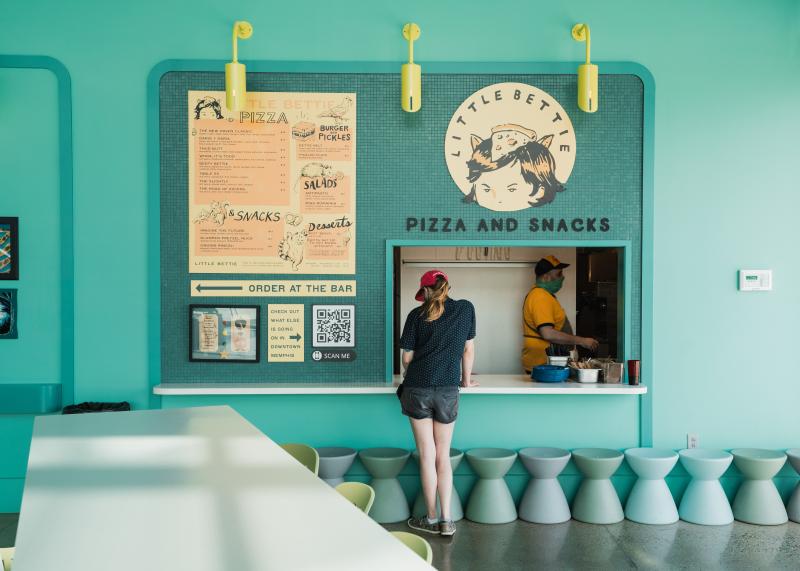 woman stands at window of little bettie food stand inside wiseacre -- walls are all teal