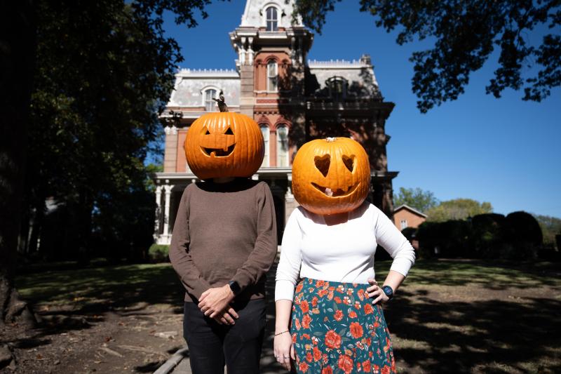 two people with pumpkin heads stand in front of victorian home