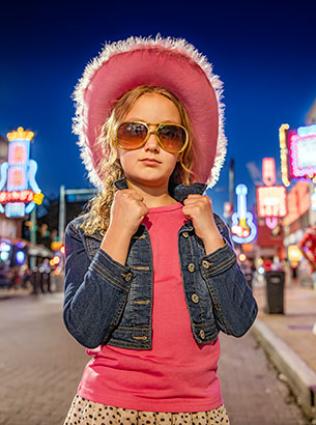 A young girl stands on neon-lit Beale Street in Memphis wearing a pink hat and Elvis Presley-inspired sunglasses