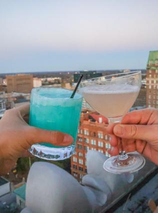 Two hands holding cocktails together in a cheers motion with a view of Downtown Memphis in the background, Photo by Alex Shansky