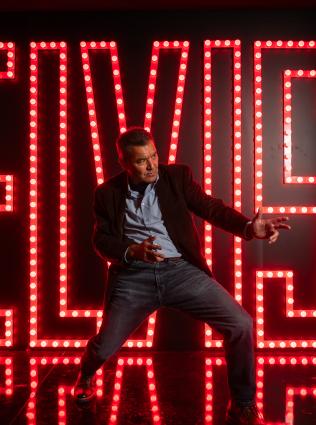 A man posing in front of the red neon Elvis sign at Elvis Presley's Graceland.