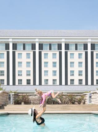 woman tosses child up in the air in the pool with guest house hotel behind them