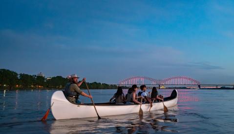 A group of people row a multi-person canoe on the Mississippi River with an illuminated bridge in the background.