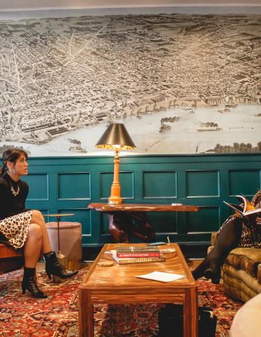 Two women sitting inside the lobby of Arrive Hotel with a large throwback illustration of the Mississippi riverfront on the wall behind them.