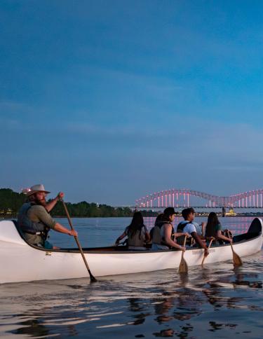 A group of people row a multi-person canoe on the Mississippi River with an illuminated bridge in the background.
