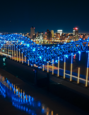 memphis "m" bridge lit up blue at night with city skyline in background