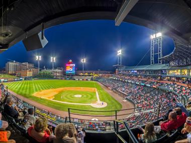 Panoramic photo of baseball stadium full of fans in the evening. The diamond is lit up by the lights