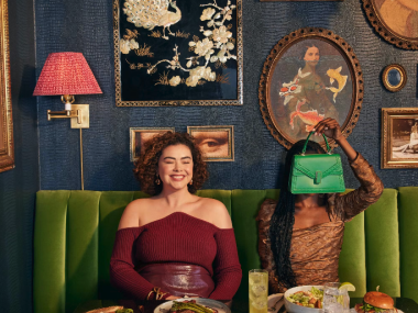 two laides sitting in front of a table full of drinks and food, one is holding a green purse up in front of her face