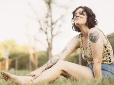 white woman with short brown hair in a yellow top and jean shorts sitting on the ground in the grass leaning forward and smiling