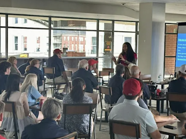 people sitting and listening while two people stand at the front of the room talking