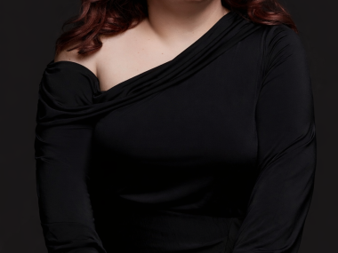 white woman with long brown hair, dark eyebrows, red lipsticks, and white flower earrings smiles softly at the camera