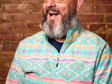 white man with very full beard and glasses smiling and looking up in front of a brick wall (picture is only from the neck up)
