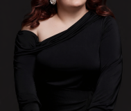 white woman with long brown hair, dark eyebrows, red lipsticks, and white flower earrings smiles softly at the camera