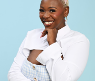 African American Woman with Blonde Buzed Hair, pink bow, smiling into the camera against a light blue background