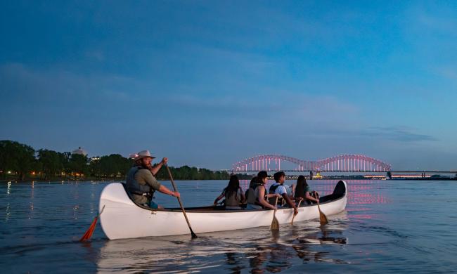 A group of people row a multi-person canoe on the Mississippi River with an illuminated bridge in the background.