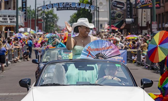 Person waves from convertible vehicle during Pride parade