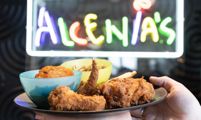 A plate of fried chicken and Southern sides with the multi-color Alcenia's neon sign lit behind it.