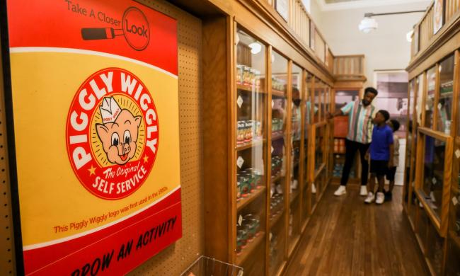 Piggly Wiggly sign in the foreground with a man and two children looking at the display shopping aisles inside the Piggly Wiggly exhibit at Pink Palace Museum &amp; Mansion.