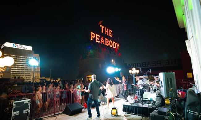 A band playing in front of a crowd at a Peabody Rooftop Party, with the red Neon "The Peabody" sign lit in the background of the crowd.