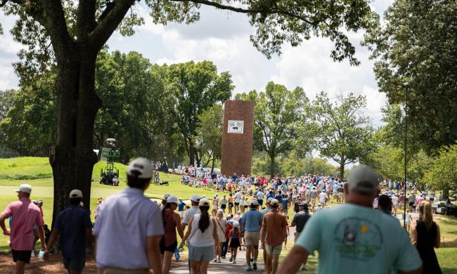 People walk along path lining golf course at FedEx St. Jude Championship with old smokestack ahead