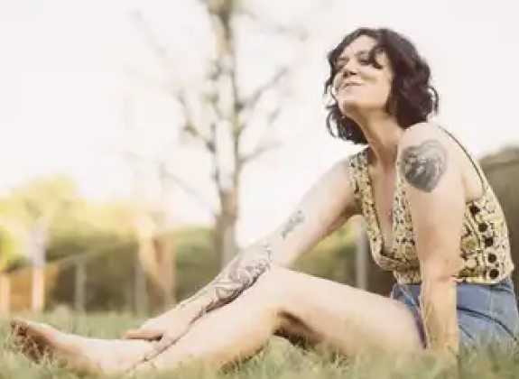 white woman with short brown hair in a yellow top and jean shorts sitting on the ground in the grass leaning forward and smiling