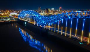 memphis "M" bridge lit up blue at night with city skyline in background