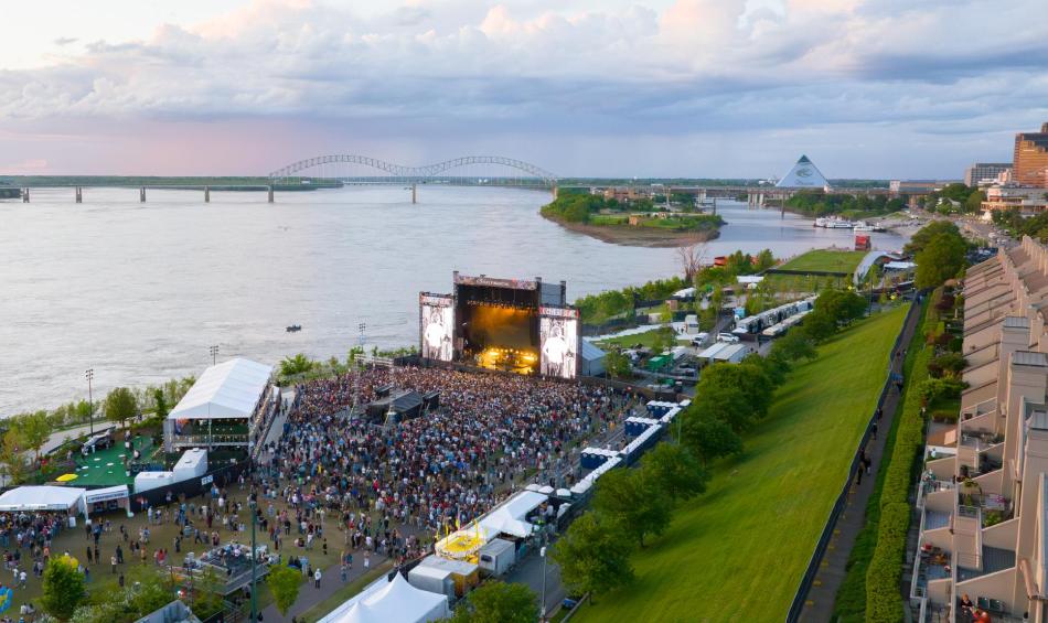 An aerial view of RiverBeat Music Fest, with a large crowd gathered in front of a large stage. The Mississippi River, Hernando de Soto Bridge and Memphis skyline are in the background.