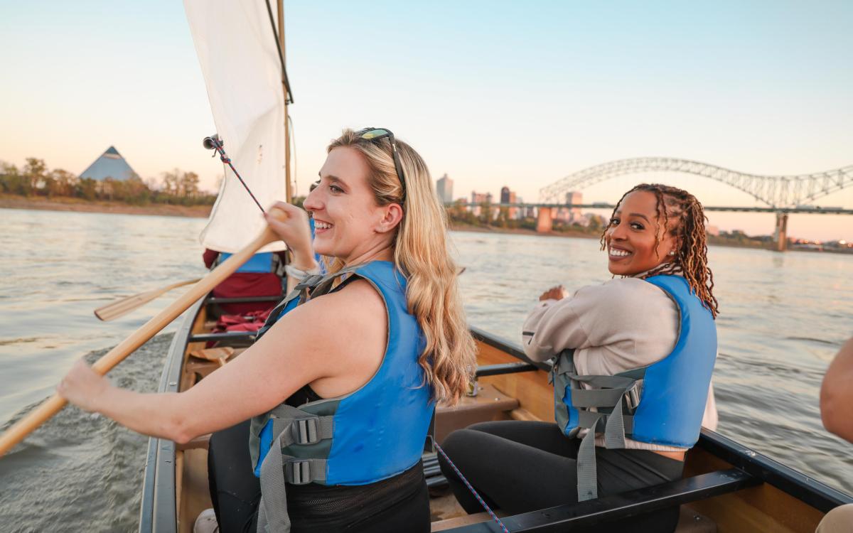 two women row canoe on Mississippi River with Memphis skyline in background 