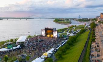 An aerial view of RiverBeat Music Fest, with a large crowd gathered in front of a large stage. The Mississippi River, Hernando de Soto Bridge and Memphis skyline are in the background.